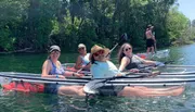 A group of smiling people are enjoying kayaking and paddleboarding on a sunny day in clear water with lush greenery in the background.