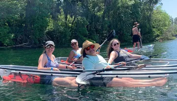 A group of smiling people are enjoying kayaking and paddleboarding on a sunny day in clear water with lush greenery in the background.