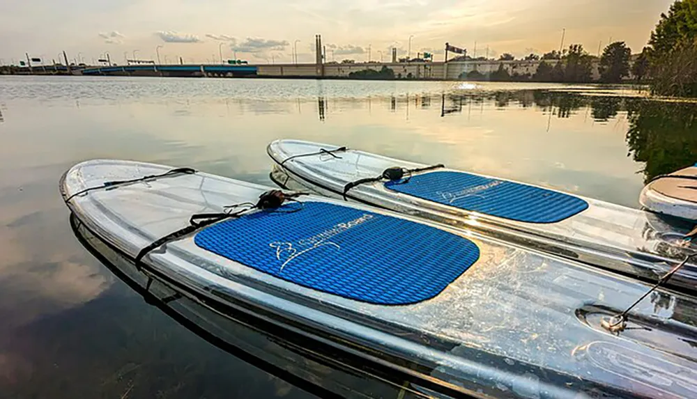 Three paddleboards are resting on a calm body of water with an industrial backdrop highlighted by the warm glow of a setting or rising sun