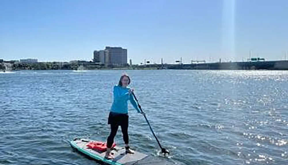 A person is paddleboarding on a body of water with a dog against a backdrop of a bright sky and distant urban buildings