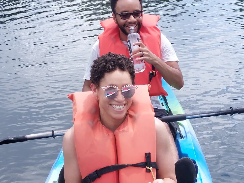 Two people are wearing orange life vests and sharing a smile while kayaking on a calm body of water