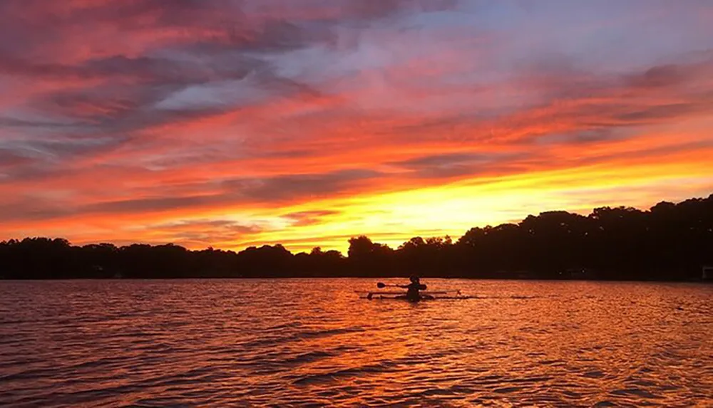 A lone rower glides across a tranquil body of water under a vibrant sunset sky painted in shades of orange and red