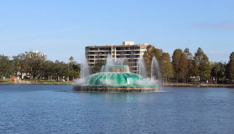 A large, tiered fountain with a turquoise-colored lower tier sprays water into the air, set against the backdrop of a lake, palm trees, and buildings under a blue sky.