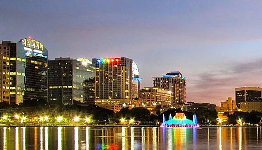 The image shows a vibrant city skyline at dusk with illuminated buildings reflecting off the water's surface, accented by a colorful lit fountain in the foreground.