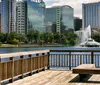 A waterfront view from a wooden deck featuring a city skyline and a fountain in the center of a lake under a blue sky