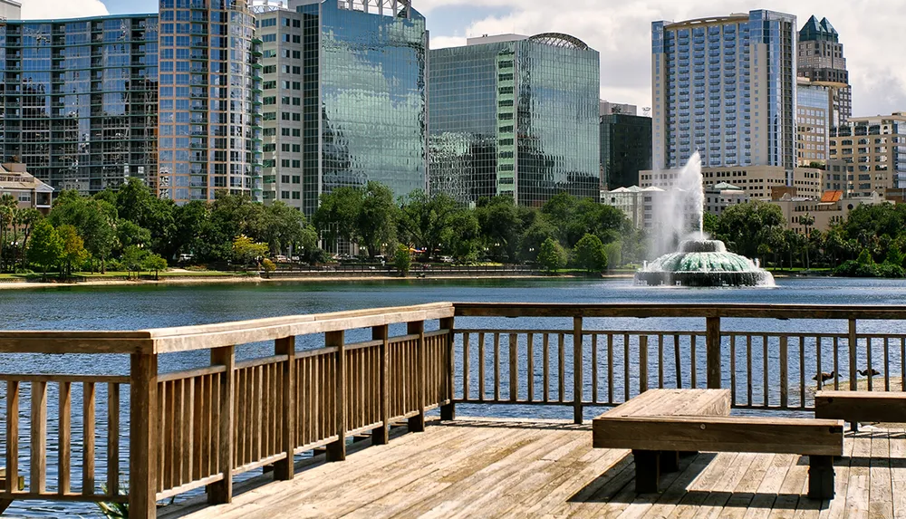 A waterfront view from a wooden deck featuring a city skyline and a fountain in the center of a lake under a blue sky