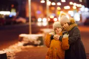 Two children warmly dressed in winter clothing are hugging affectionately on a city street lit by the glow of streetlights and storefronts at dusk or night.