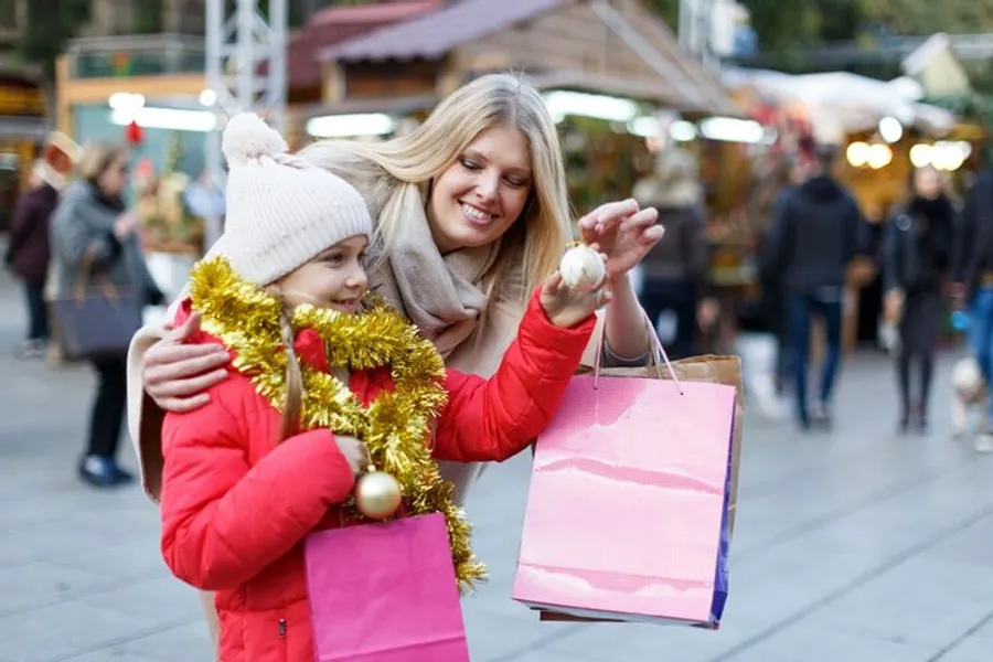 A smiling woman and a young girl, who is holding a golden tinsel garland and a shopping bag, appear to be enjoying a festive outdoor holiday market.