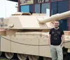 A man is smiling for a photo in front of a large tan-colored military tank displayed outdoors