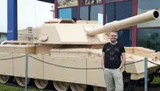 A man is smiling for a photo in front of a large, tan-colored military tank displayed outdoors.