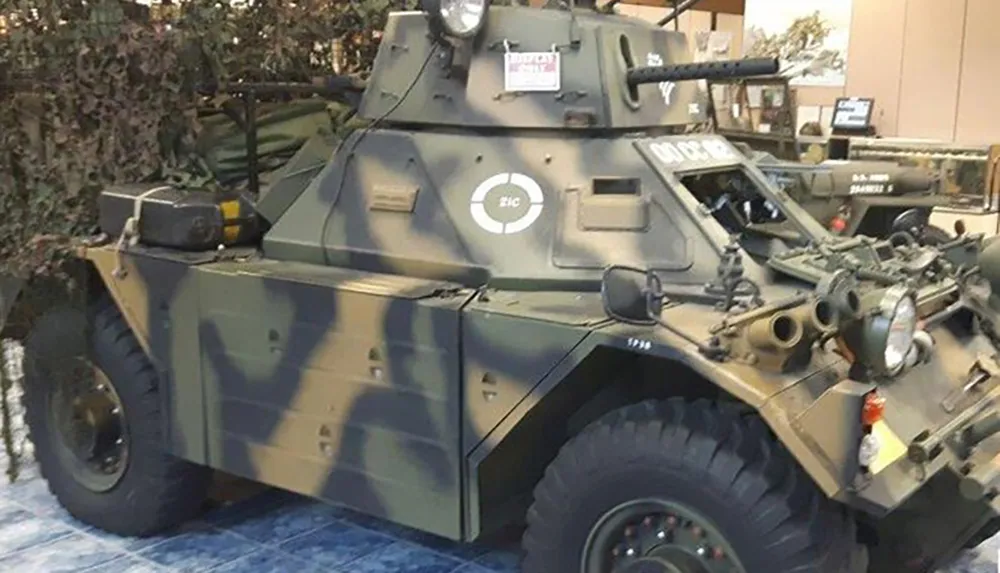 The image shows a camouflage painted military armored vehicle equipped with a turret and multiple antennas and it appears to be on display with a blurred backdrop that may suggest an exhibition setting