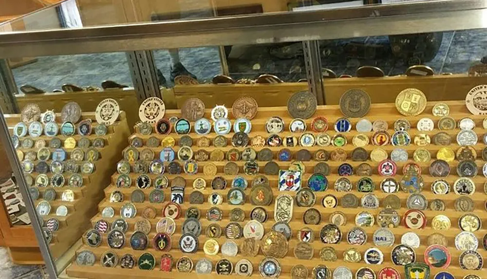 The image shows a collection of various colorful challenge coins displayed on tiered shelves in a glass case