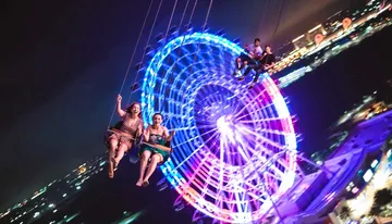 People are enjoying a nighttime swing ride against the backdrop of a brightly lit Ferris wheel and city lights.