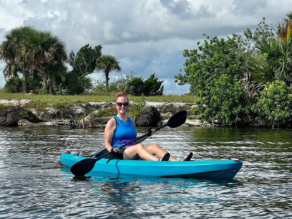A person is smiling while seated in a blue kayak on a calm water body with tropical vegetation in the background