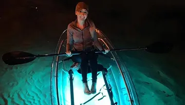 A person is kayaking at night in a clear kayak illuminated from within, highlighting the water below.