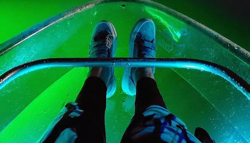 A person stands on a clear kayak or boat revealing an illuminated underwater view glowing in green