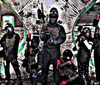 Two people wearing masks and protective vests are holding paintball guns standing next to an inflatable barrier in a paintball arena