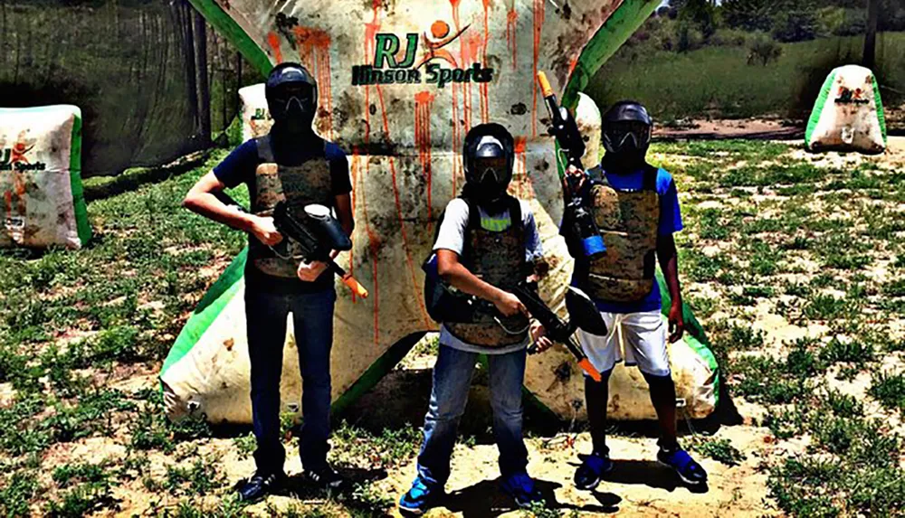 Three people in protective gear and masks holding paintball guns are posing in front of a paint-splattered inflatable barricade on a paintball field