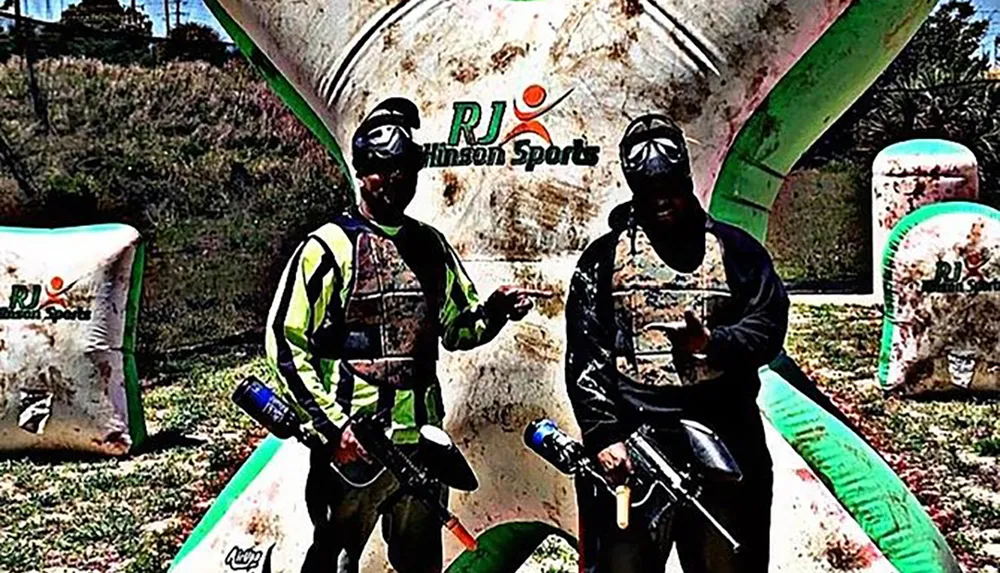 Two people wearing protective gear and holding paintball guns are posing in front of inflatable barriers at a paintball facility