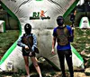 Two people wearing masks and protective vests are holding paintball guns standing next to an inflatable barrier in a paintball arena
