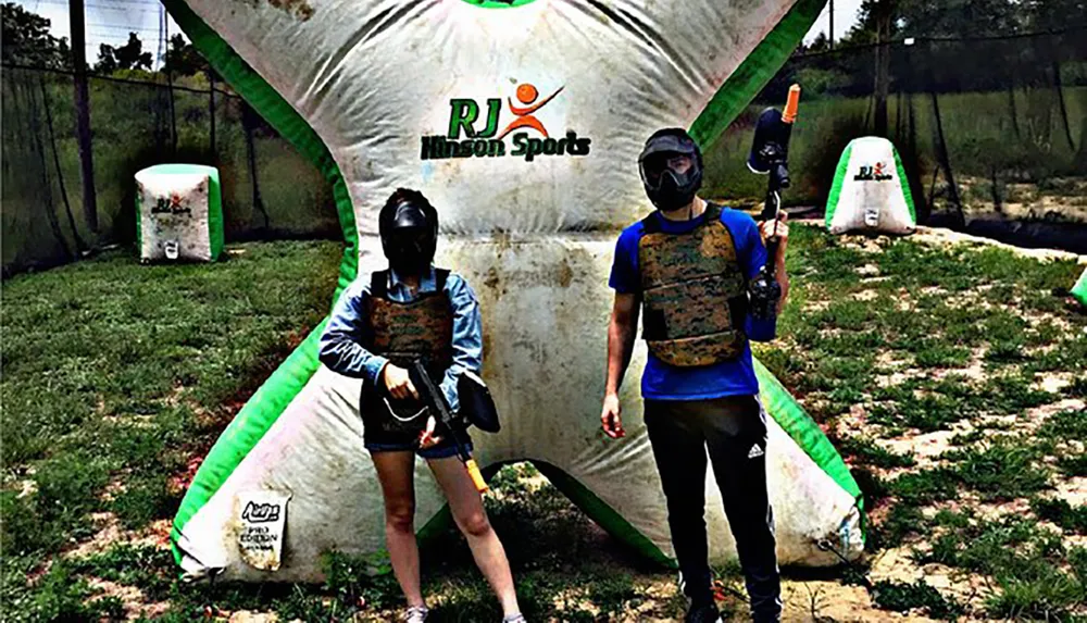 Two people wearing masks and protective vests are holding paintball guns standing next to an inflatable barrier in a paintball arena