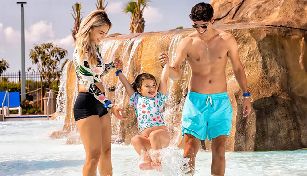 Two adults and a child are enjoying themselves in a water park with smiles all around as they play in the water near an artificial waterfall
