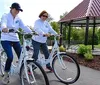 Two individuals are smiling and riding matching white bicycles on a paved path near a gazebo in a park-like setting