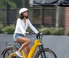 Two individuals are smiling and riding matching white bicycles on a paved path near a gazebo in a park-like setting