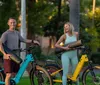 Two individuals are smiling and riding matching white bicycles on a paved path near a gazebo in a park-like setting