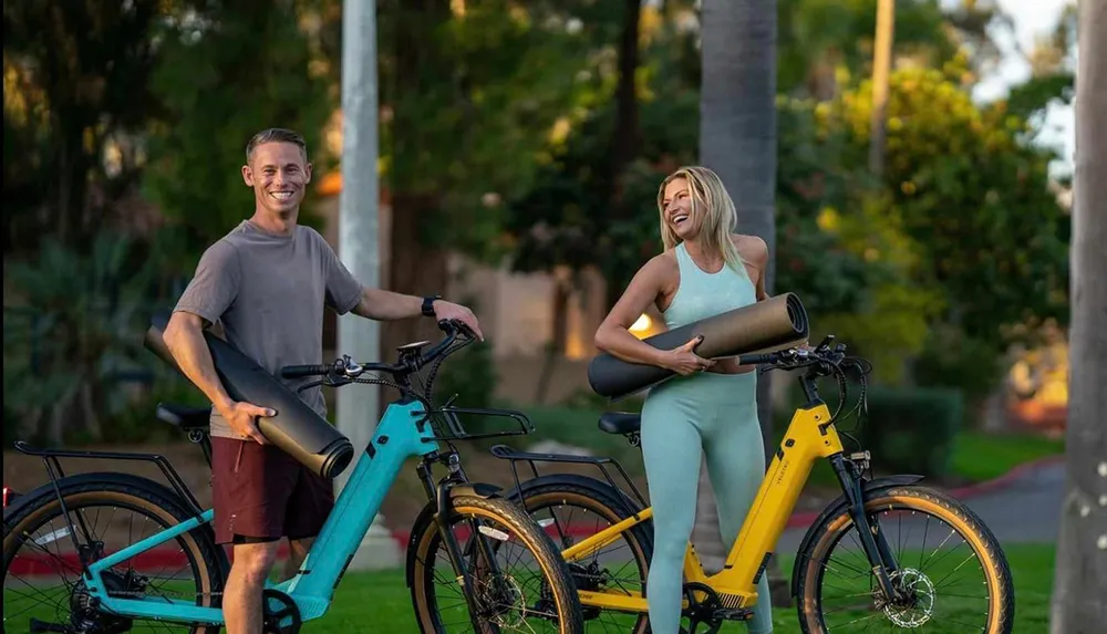 A man and a woman are smiling and holding exercise equipment next to two colorful bicycles in a park-like setting