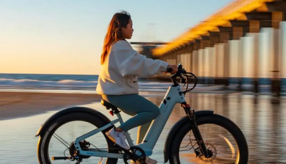 A person is riding a bicycle along a beachside path at sunset with a blurred pier in the background creating a motion effect