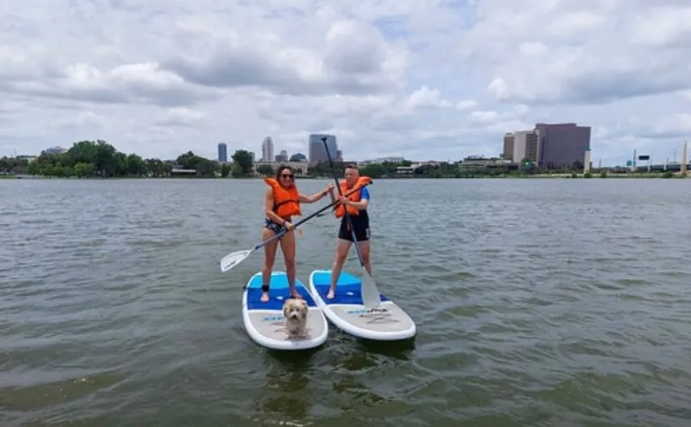 Two people and a dog are paddleboarding on a body of water with a city skyline in the background