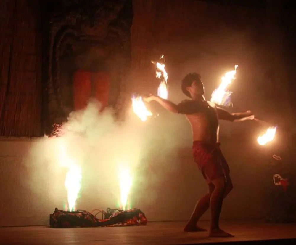A performer is seen executing a fire dance with lit torches in each hand against an illuminated foggy background on stage