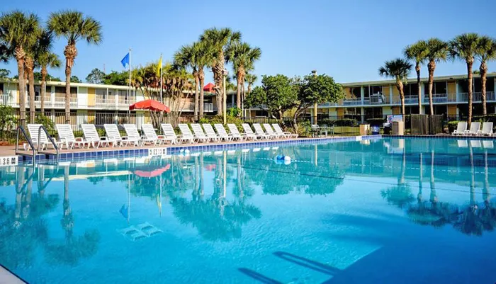 A clear sunny day at an outdoor swimming pool with rows of lounge chairs and palm trees reflecting on the water adjacent to a multi-story building
