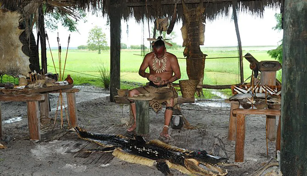 An individual wearing indigenous attire is sitting under a thatched structure amidst cultural artifacts and a crafted alligator display