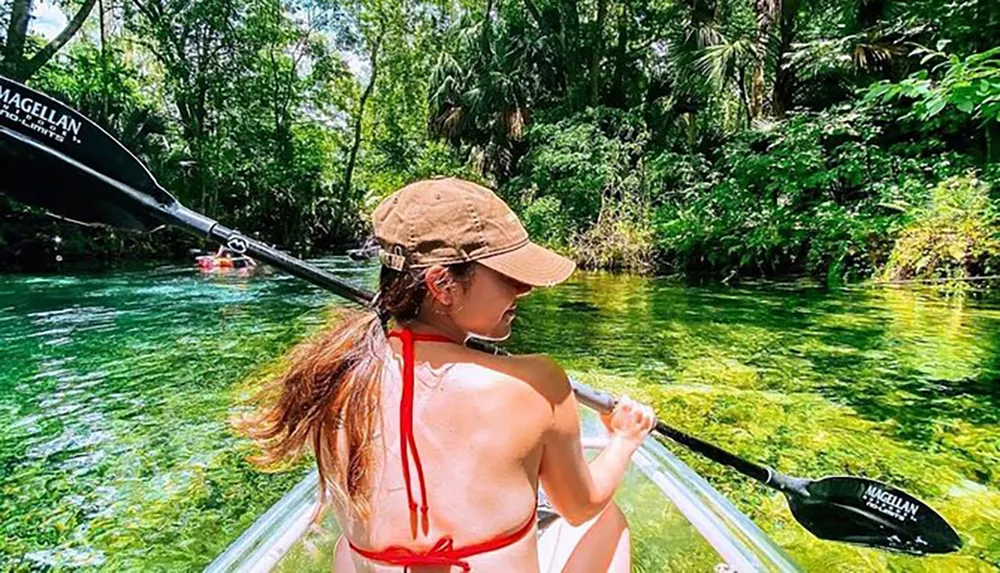 A person is kayaking in clear verdant waters surrounded by lush greenery on a sunny day