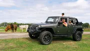 A family enjoys a drive in a black Jeep Wrangler with cows in the background in a countrified setting.