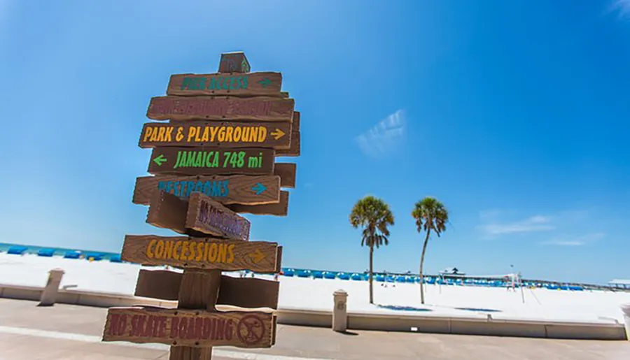 A wooden direction signpost with multiple arrows points to various amenities and destinations against a backdrop of palm trees and a clear blue sky at a sunny beachside location.