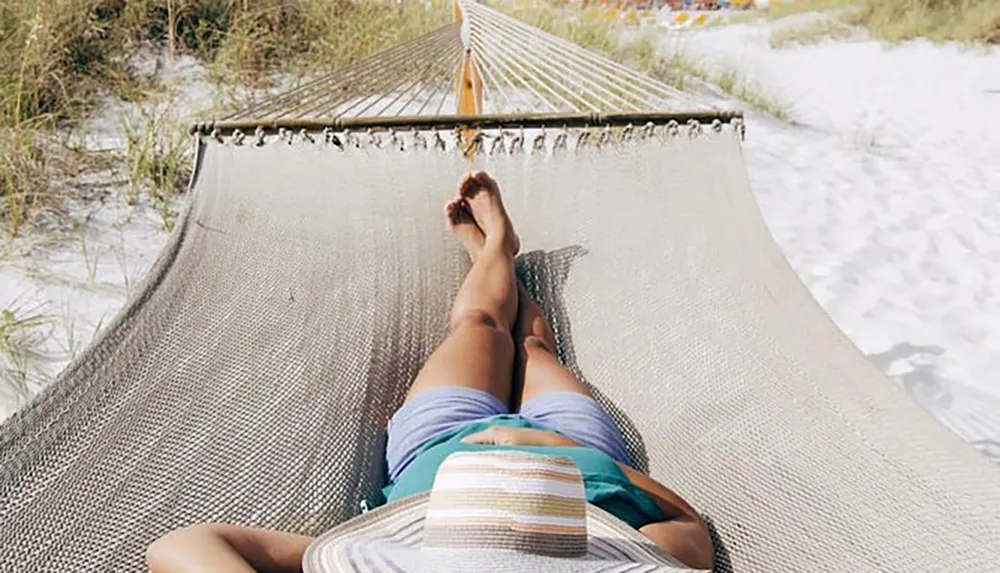 A person relaxes on a hammock by the beach their view pointing towards their feet with sand and beach vegetation in the background