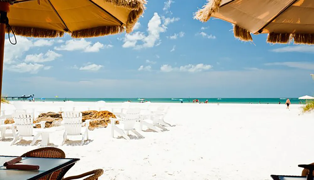 This image shows a sunny beach scene viewed from under a thatched umbrella with white Adirondack chairs on the sand leading to a calm blue sea and a clear sky with a few scattered clouds