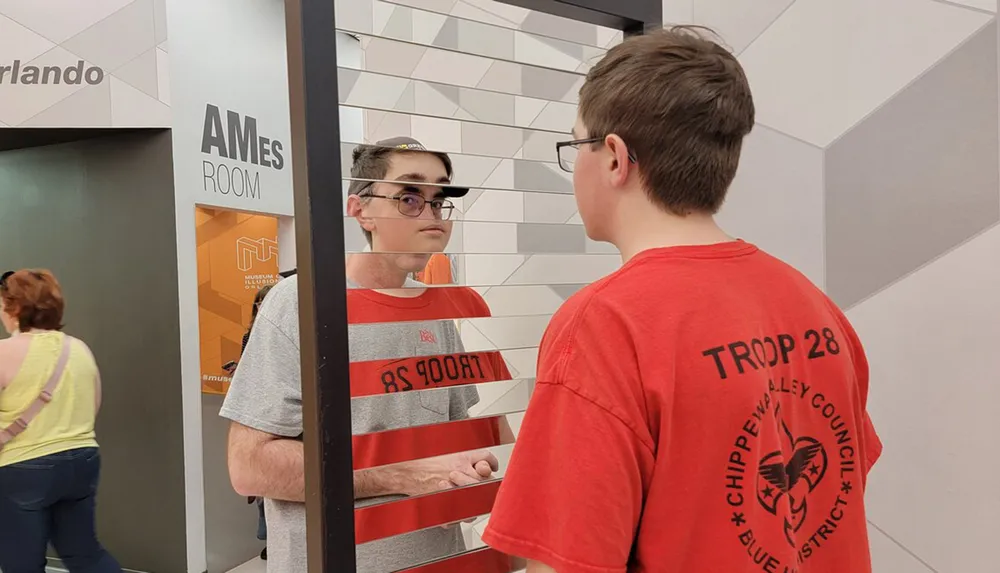 Two people are interacting near an Ames Room exhibit with one appearing disproportionately large due to the optical illusion created by the room