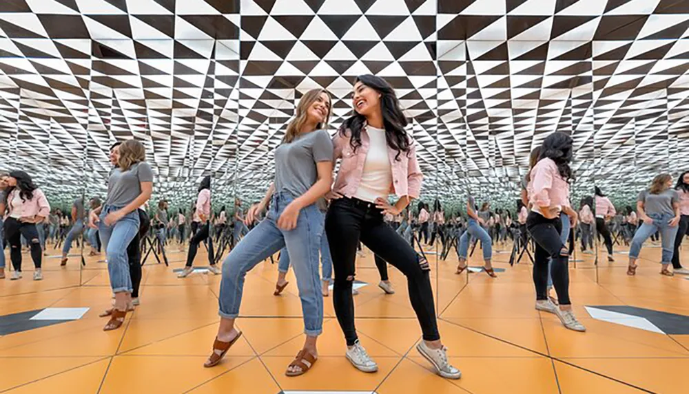 Two women are laughing and posing for a photo in a mirrored room that creates an infinite reflection effect