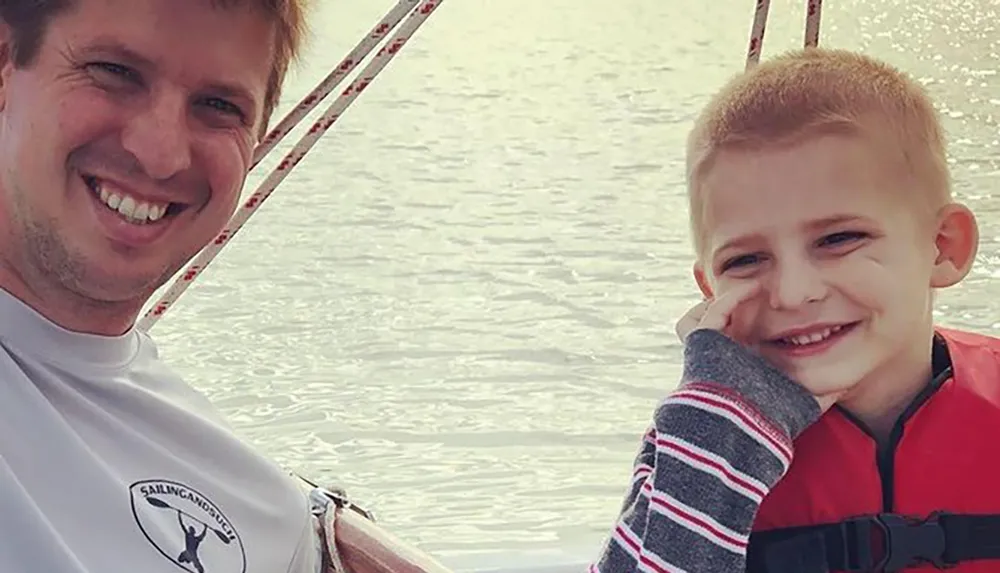 A man and a young boy are smiling at the camera with a backdrop of water possibly out on a boat