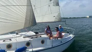 Three people are enjoying a sunny day sailing on a white sailboat with the coastline in the background.