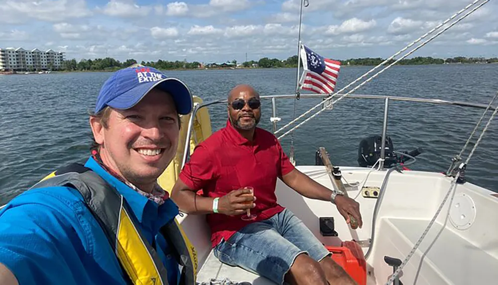 Two men are smiling and enjoying a sunny day on a sailboat with one of them taking a selfie and an American flag visible in the background