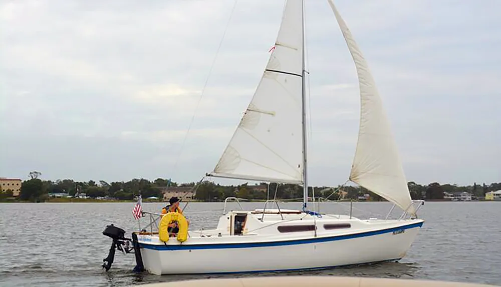 A person in a yellow life jacket is sitting on a small sailboat with sails up floating near the shore on a calm body of water