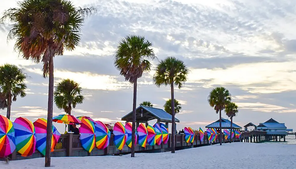 A row of vibrant multicolored beach umbrellas lines the sandy shore under a sky dotted with clouds flanked by palm trees and a wooden pier in the background