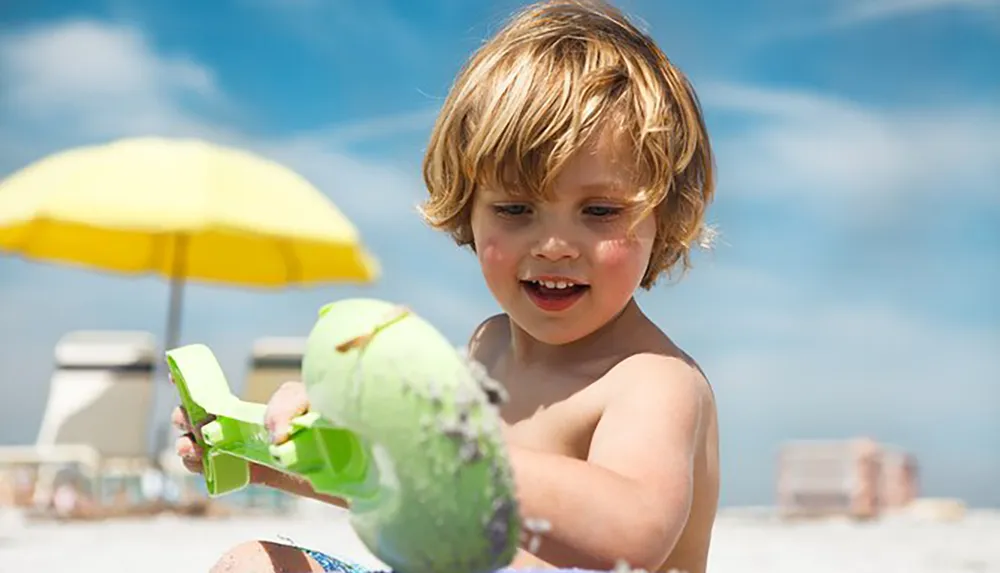 A young child is playing with a bright green toy in the sand on a sunny beach with a yellow beach umbrella in the background