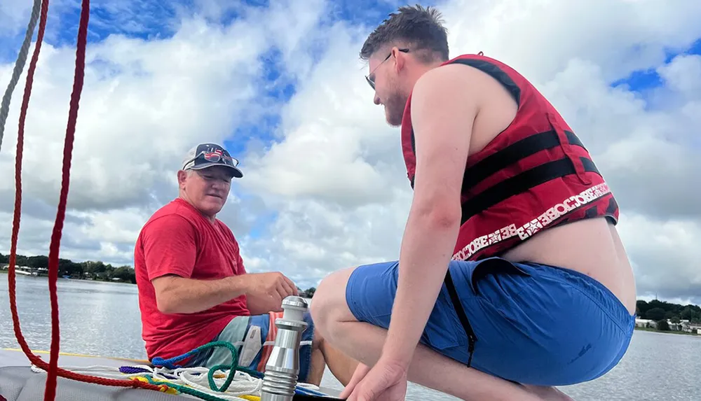 Two individuals are on a sailboat one sitting and steering while the other is kneeling and wearing a life jacket with ropes and a body of water in the background