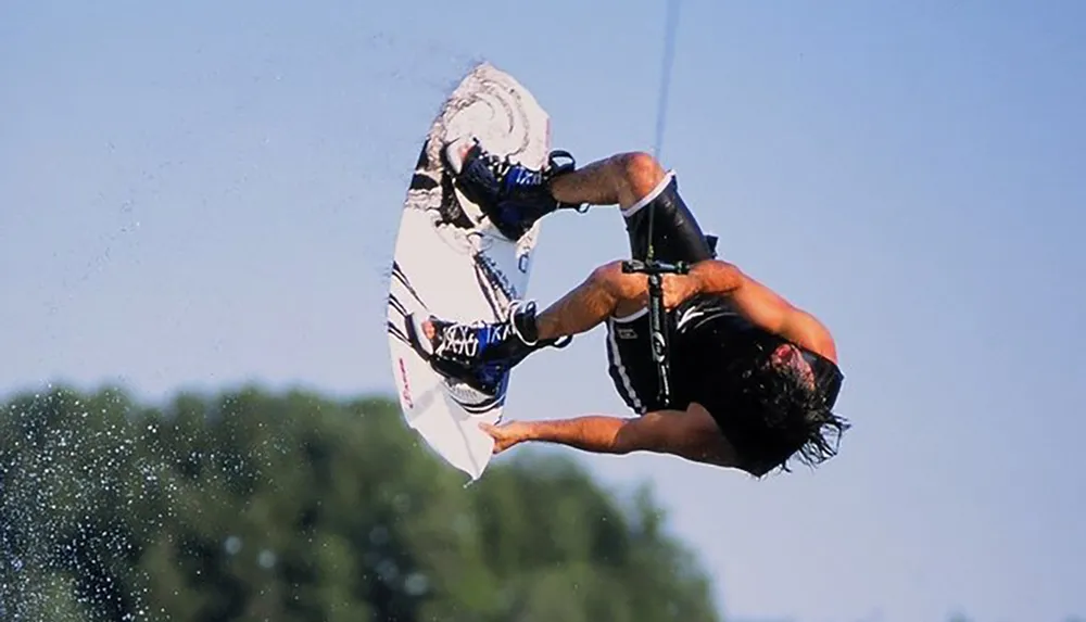 A wakeboarder is captured midair performing an upside-down trick while holding onto a towrope against a clear blue sky